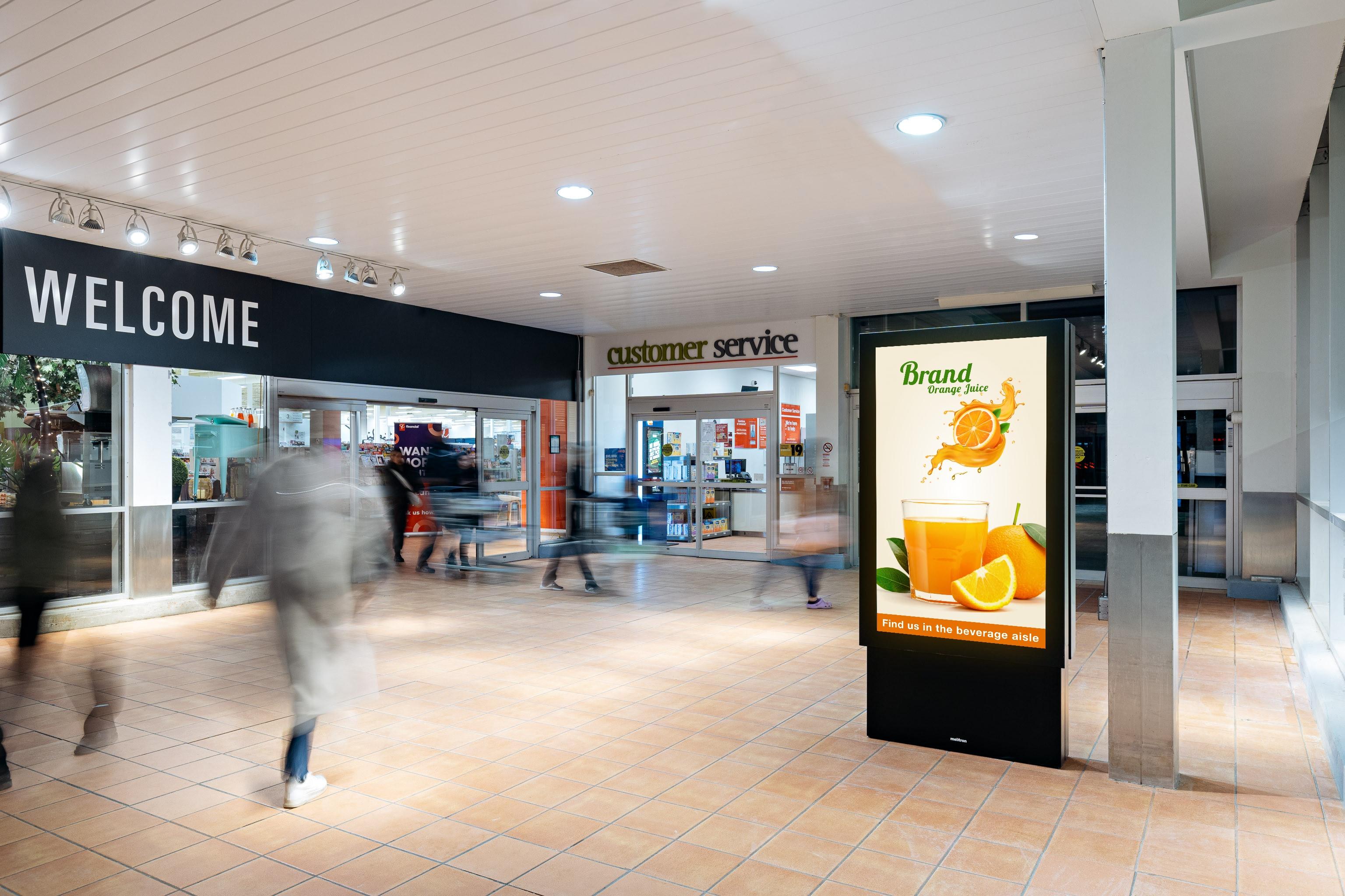 A screen at the front entrance of a grocery store with an advertisement for orange juice on it.